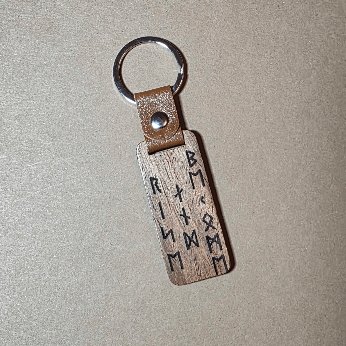 Walnut and leather Viking keychain with runic inscription “Rise and Become” lying on a neutral background